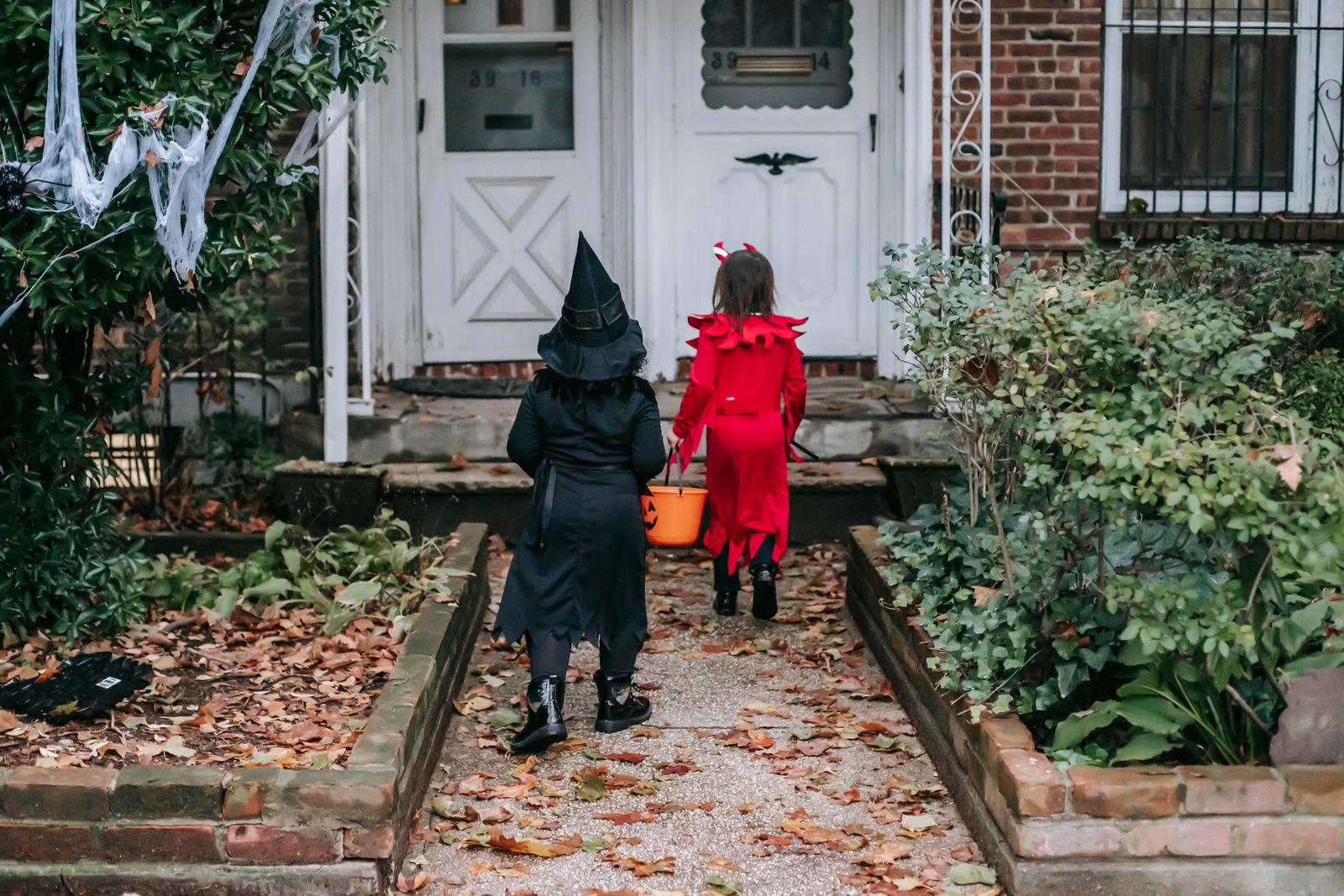 Two children in witch and devil Halloween costumes carrying a jack-o-lantern candy bucket up to a front door